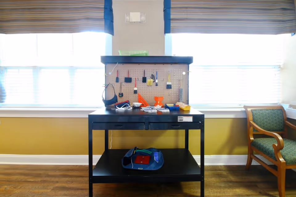A small black workbench holding tools and supplies sits in front of bright windows in a sitting area with a wooden chair.