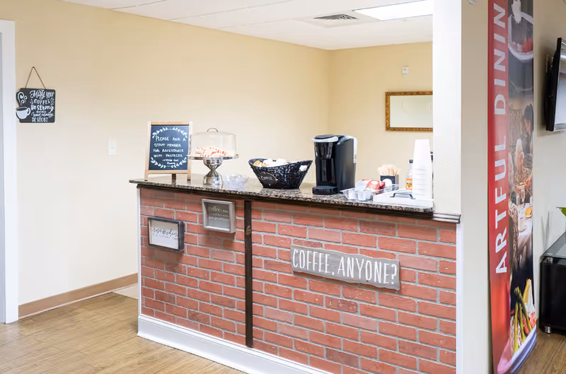 A small brick-front coffee counter with a Keurig, cups, snacks and a sign that says "Coffee, anyone?" in a communal dining area.