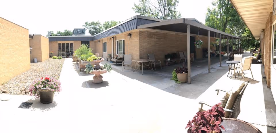 Outdoor patio area at Briarwood Health Care Center with brick walls, several potted plants, and multiple seating arrangements including chairs and small tables under a covered walkway and in open sunlight.