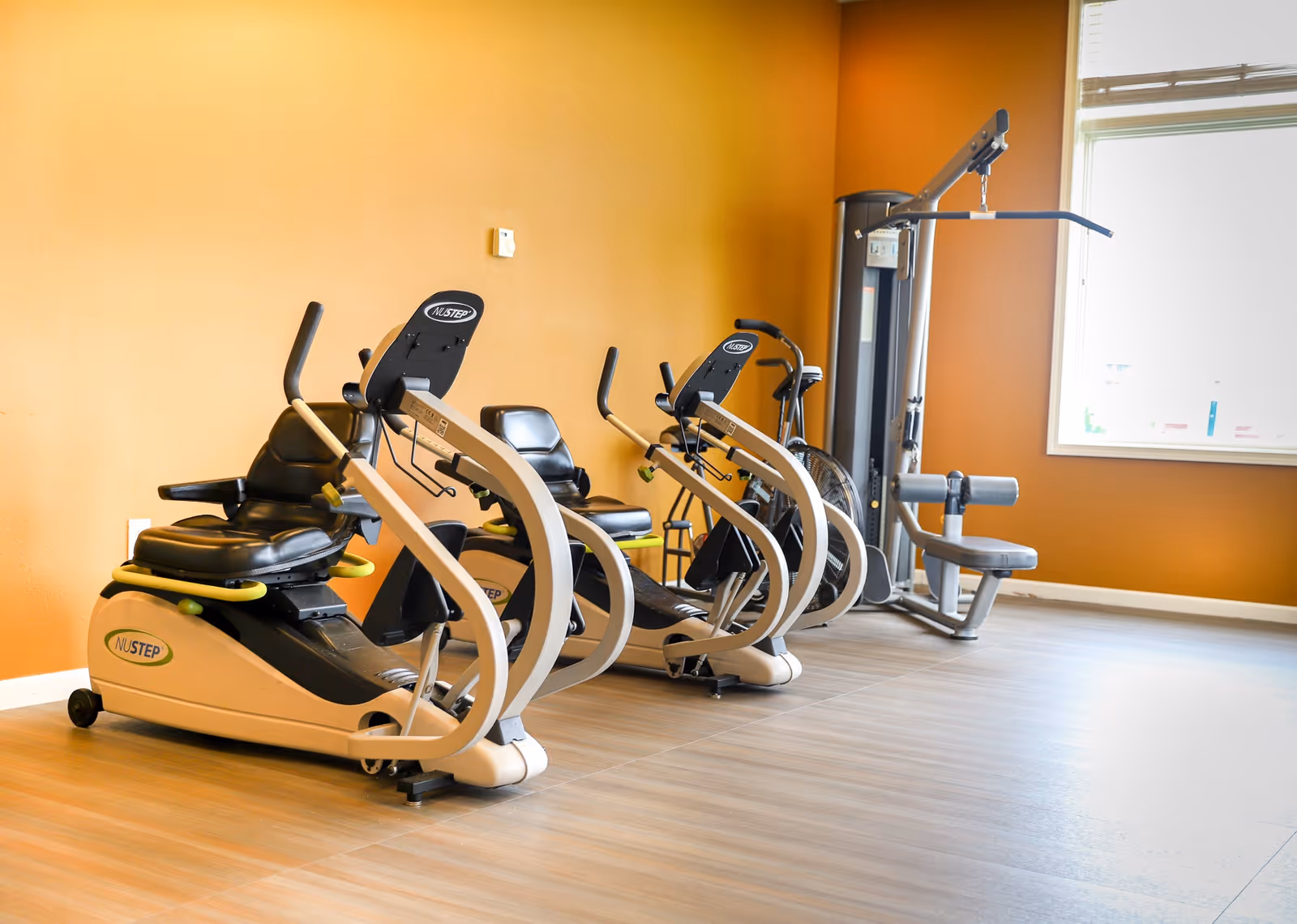 An exercise room with recumbent stationary bikes and a weight machine against an orange wall and wood-look floor.