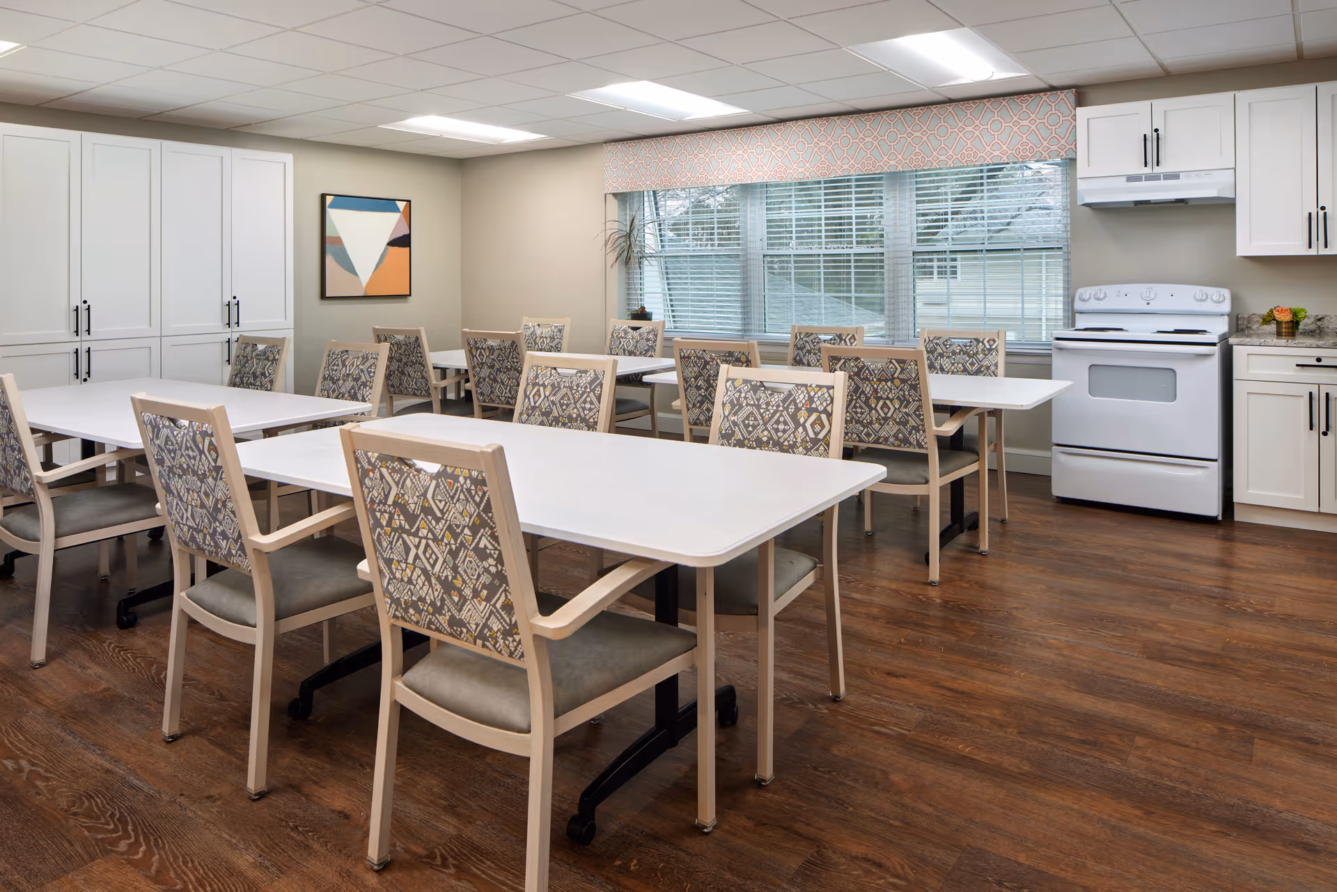 Bright dining/activity room with multiple white tables and patterned chairs beside a small kitchen and large windows.