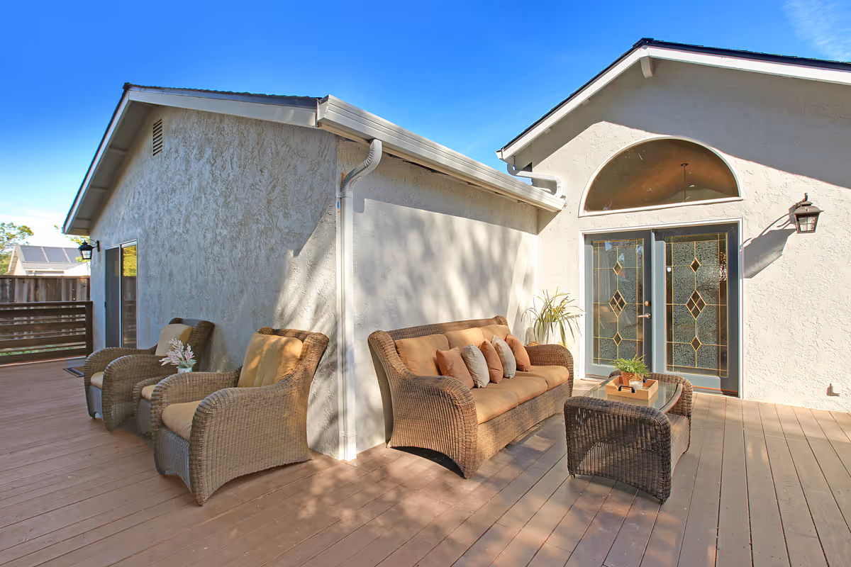 Outdoor patio area with wicker furniture including a sofa with multiple cushions, two armchairs, and a small table with a plant. The patio is attached to a light-colored building with decorative glass double doors and a large arched window above. The sky is clear and blue.