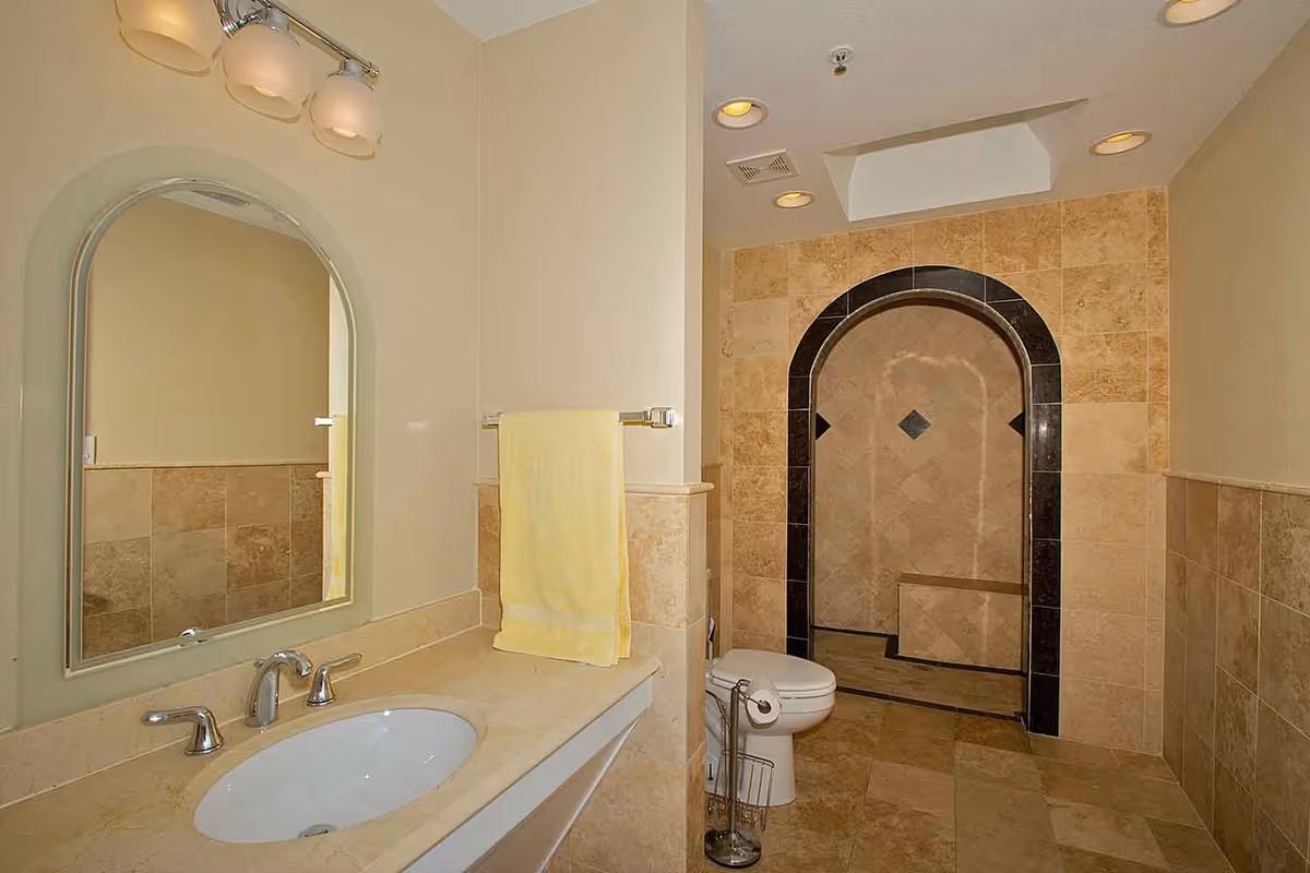 A bathroom with beige tiled walls and floor, featuring a white sink with a marble countertop, a large arched mirror above the sink, a yellow towel hanging on a towel rack, a toilet, and a walk-in shower with an arched entrance framed by dark tiles.