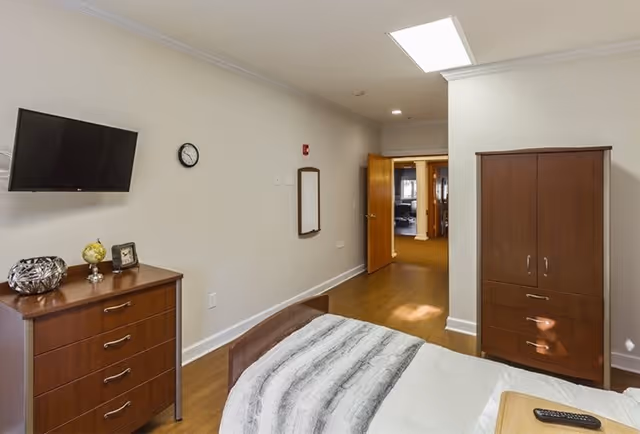 A senior living facility bedroom with a bed covered in a white and gray blanket, a wooden dresser with decorative items, a wall-mounted flat screen TV, a clock, and a wooden wardrobe. The room has light-colored walls and wood flooring, with an open door leading to a hallway.