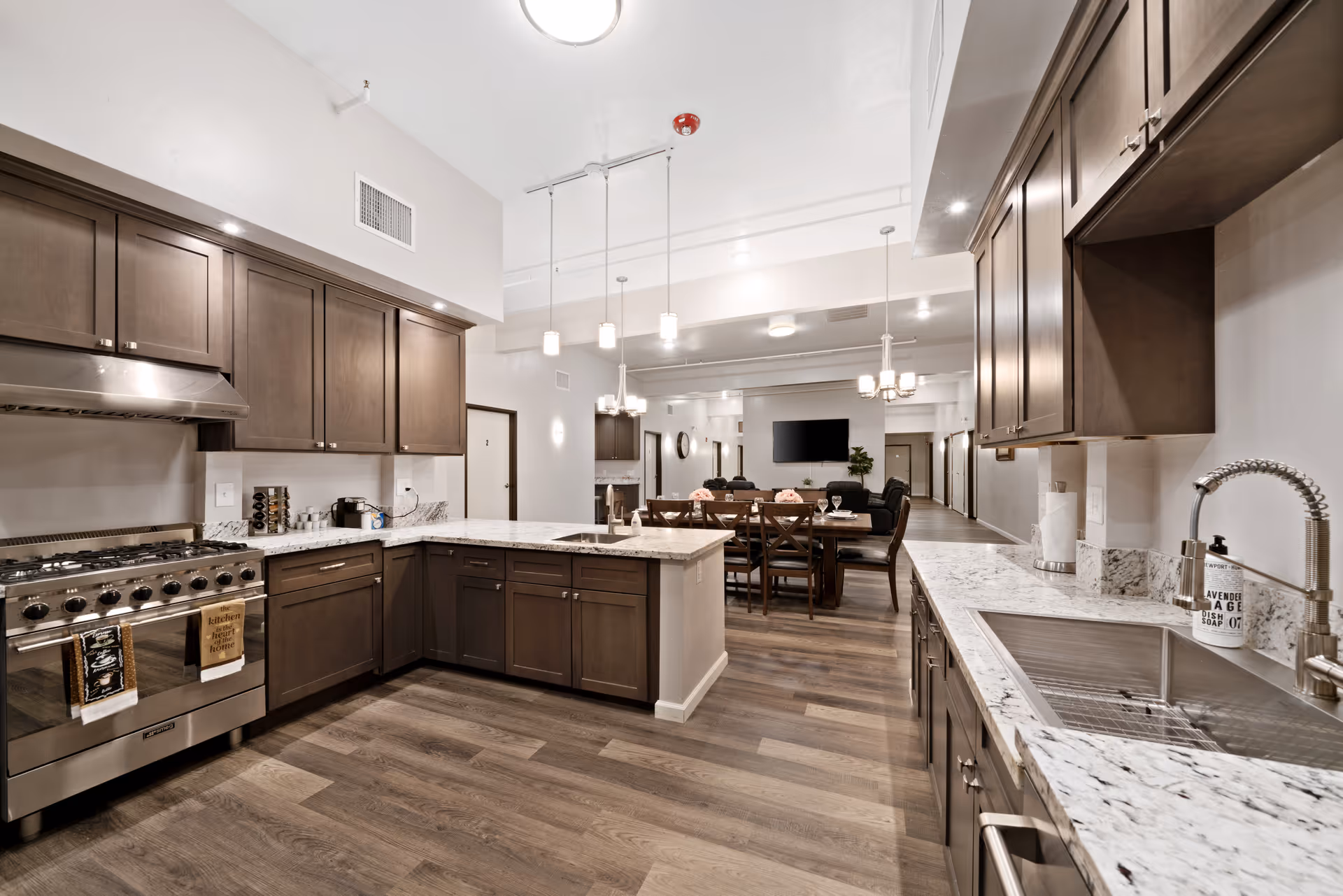 A modern kitchen with dark wood cabinets, white marble countertops, and stainless steel appliances including a large stove and a sink with a flexible faucet. The kitchen opens into a dining area with a wooden table and chairs, and a living area with a wall-mounted TV and comfortable seating. The space features wood flooring and pendant lighting.