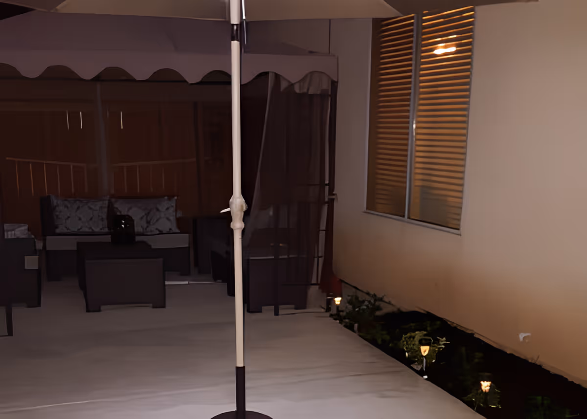Outdoor patio area at night with a canopy tent covering cushioned seating and a table. There is a large umbrella pole in the foreground and a window with blinds on the right side. Small garden lights illuminate plants along the edge of the patio.