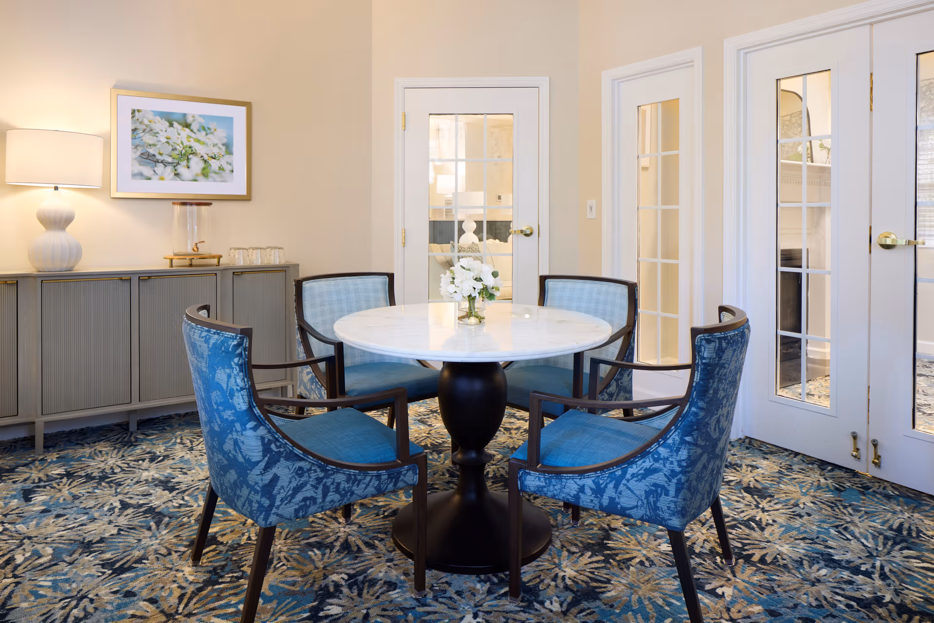 A small round dining table with a white marble top and a black pedestal base surrounded by four blue upholstered chairs with wooden frames. The room has a patterned blue carpet, beige walls, and white French doors with glass panes. A sideboard with a lamp, a framed floral picture, and a water dispenser with glasses is against the wall.