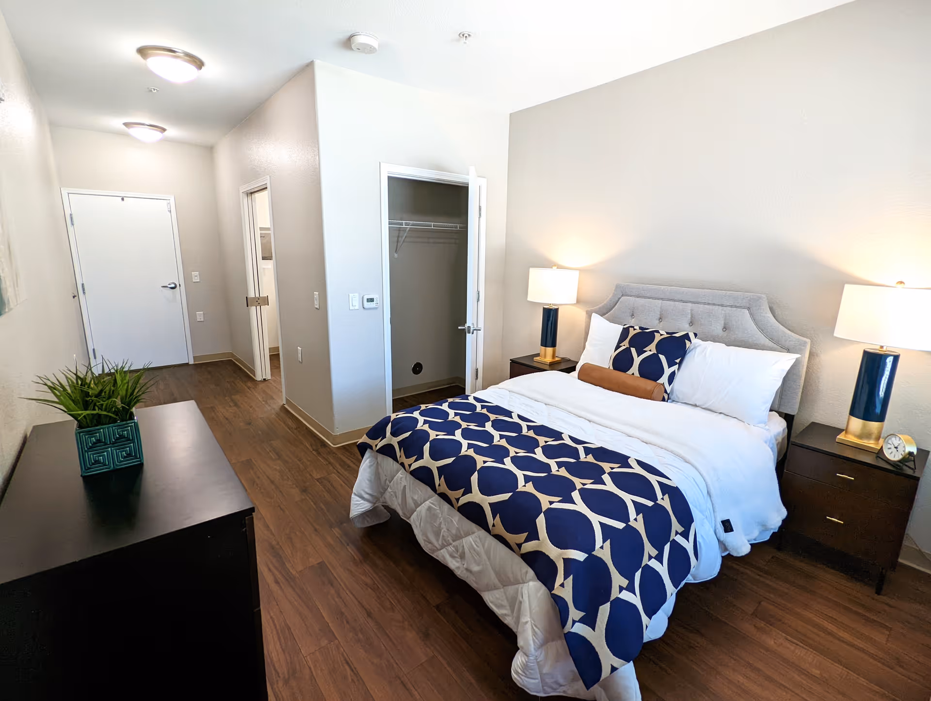 A neatly made bed with a blue and white patterned blanket and multiple pillows in a bedroom. The room has wooden flooring, two bedside tables with matching lamps, a dresser with a green potted plant, an open closet, and a hallway leading to a door and a bathroom.