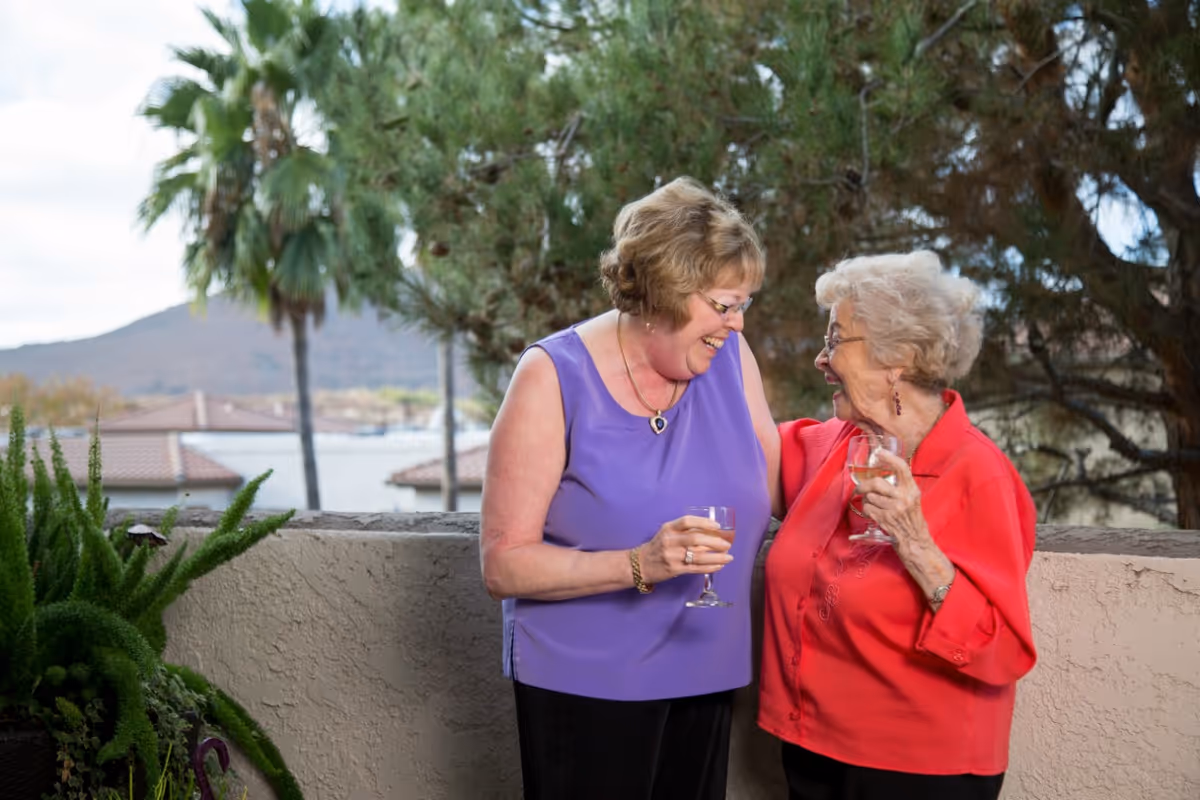 Two elderly women standing outdoors on a balcony or patio, smiling and holding glasses of wine, with trees and rooftops in the background.