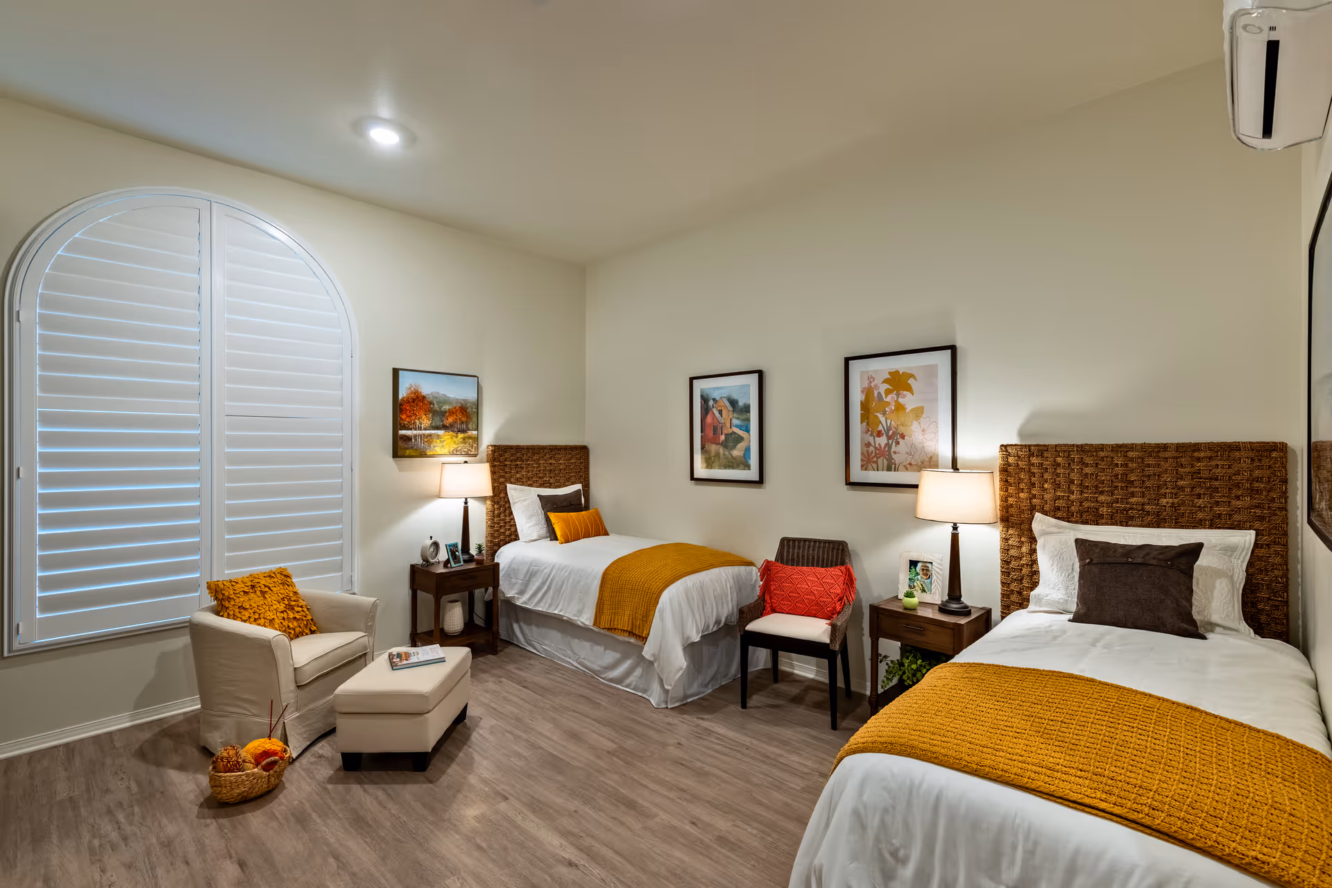 A cozy bedroom with two single beds featuring woven headboards and white bedding with mustard yellow throws. Between the beds are two wooden nightstands with lamps and decorative items. A cushioned armchair with a matching ottoman and a mustard yellow pillow is placed near a large arched window with white shutters. The walls are adorned with framed artwork, and the floor is a light wood laminate.