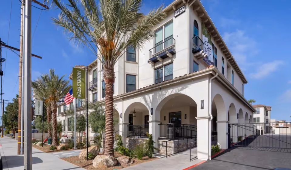 Entrance and facade of a three-story Mediterranean-style senior living building with arches, palm trees, and a gated driveway.