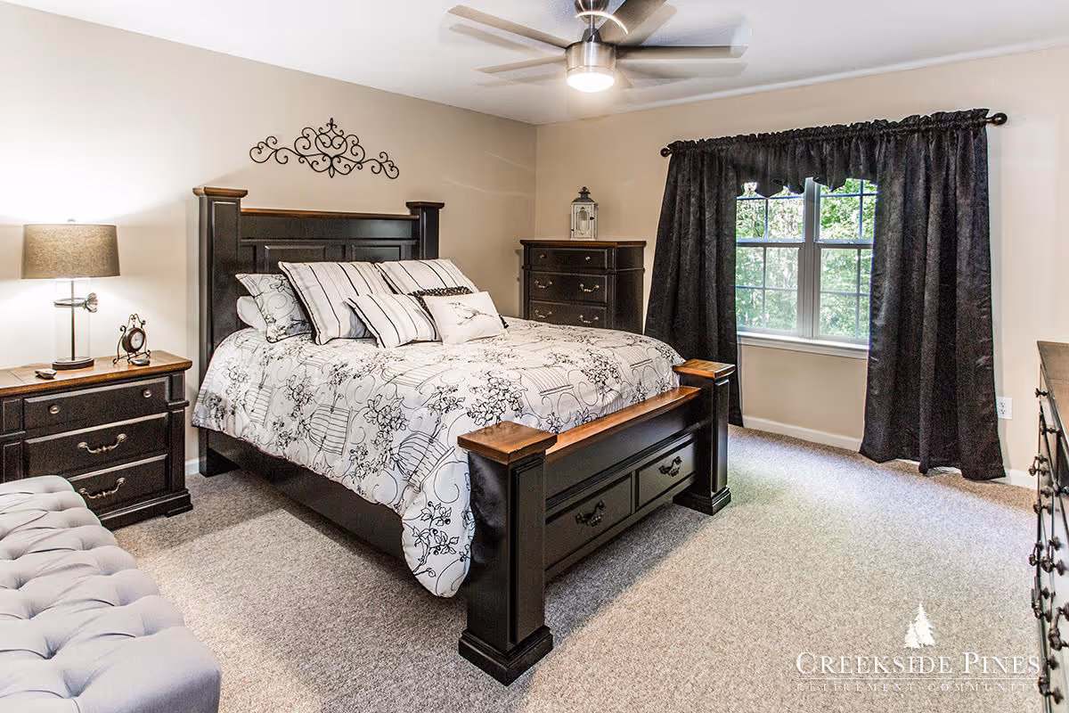 Well-lit bedroom with a large dark wood bed, matching nightstands and dresser, ceiling fan, and a window with dark curtains.