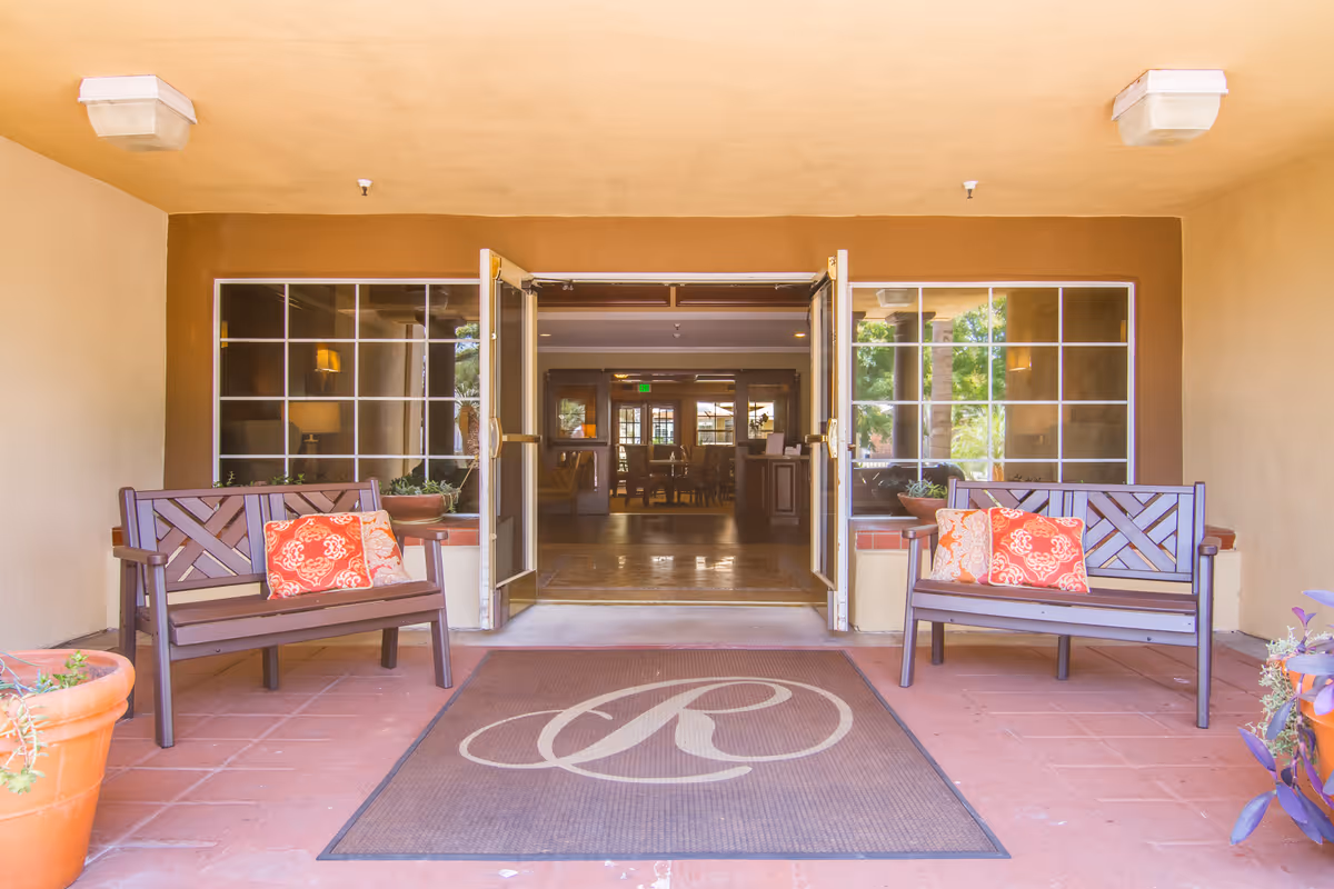 Entrance area of The Remington Club facility with two wooden benches on either side, each with decorative orange and patterned cushions. There are large windows on both sides of the double glass doors that are open, revealing an interior space with tables and chairs. A brown mat with a large stylized letter 'R' is placed in front of the entrance. Potted plants are visible on both sides of the entrance.