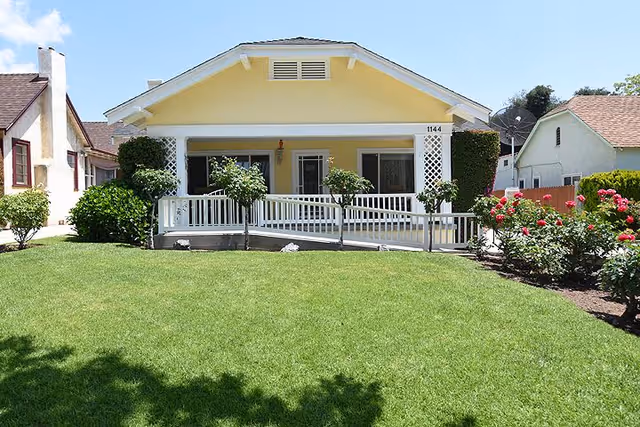 Front view of a single-story yellow house with a white porch railing and a wheelchair ramp. The house is surrounded by a well-maintained green lawn, small trees, and flowering bushes under a partly cloudy sky.