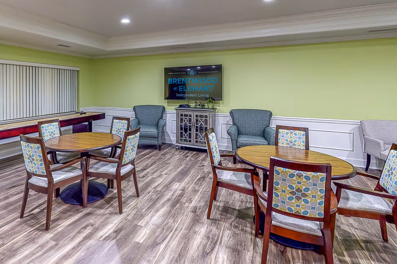A well-lit common room with two round wooden tables surrounded by chairs with patterned upholstery. There are two comfortable armchairs against a light green wall with white wainscoting. A flat-screen TV mounted on the wall displays the text 'BRENTWOOD at ELKHART Independent Living'. The floor is covered with wood-style flooring, and there is a shuffleboard table near a window with vertical blinds.