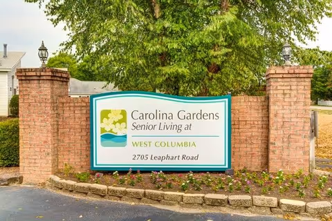 A brick sign structure with a white and teal sign that reads 'Carolina Gardens Senior Living at West Columbia, 2705 Leaphart Road' surrounded by a small landscaped area with flowers and greenery, with trees and houses in the background.