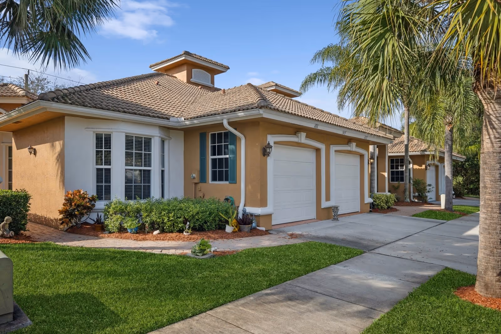 Exterior view of a single-story residential building with a tiled roof, two white garage doors, and beige walls. The front yard features green grass, palm trees, and various shrubs and plants along a paved walkway.