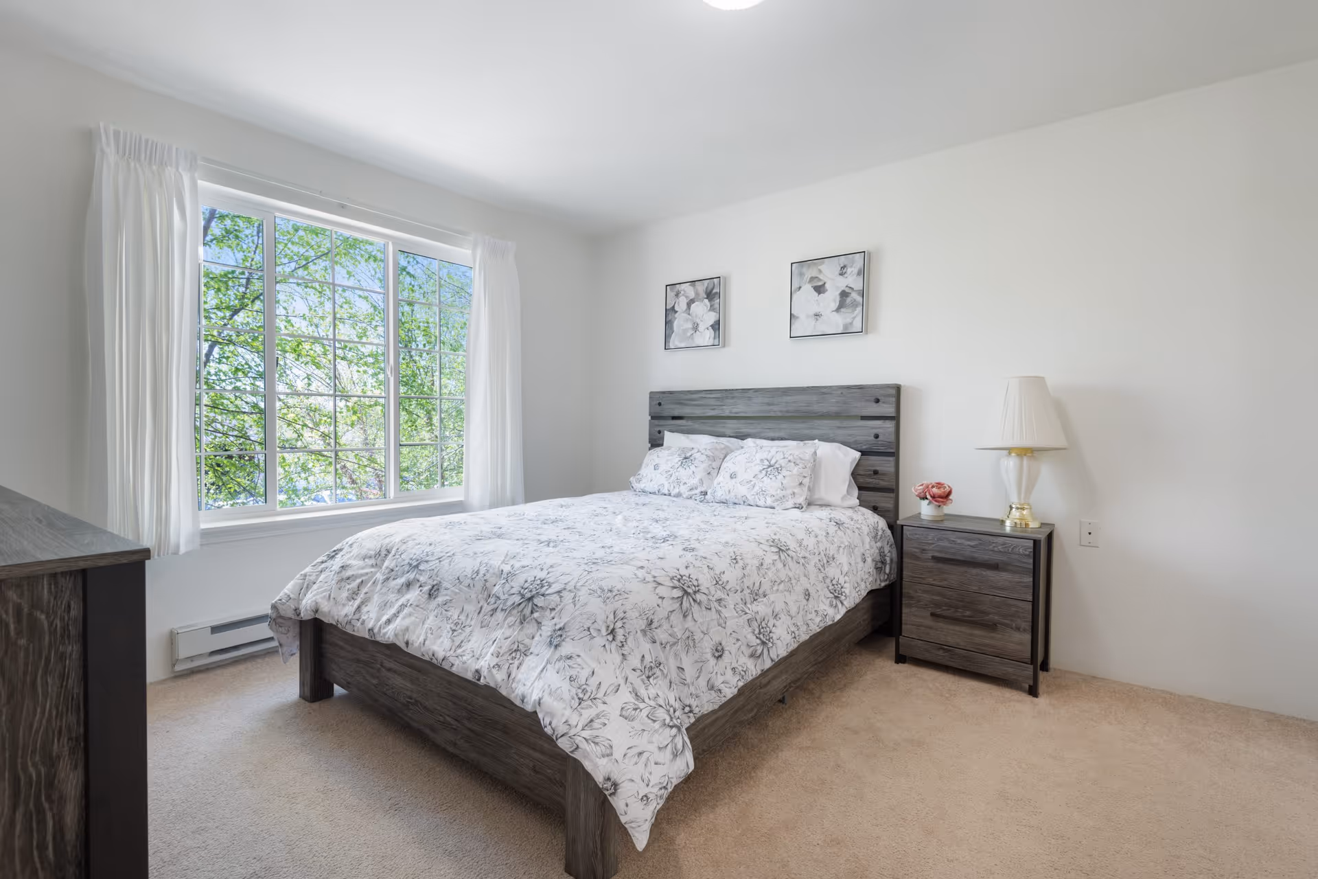 A bright bedroom with a large window showing green trees outside. The room features a wooden bed with a floral-patterned white and gray comforter and matching pillows. Next to the bed is a wooden nightstand with a white lamp and a small flower arrangement. The walls are white and the carpet is beige.