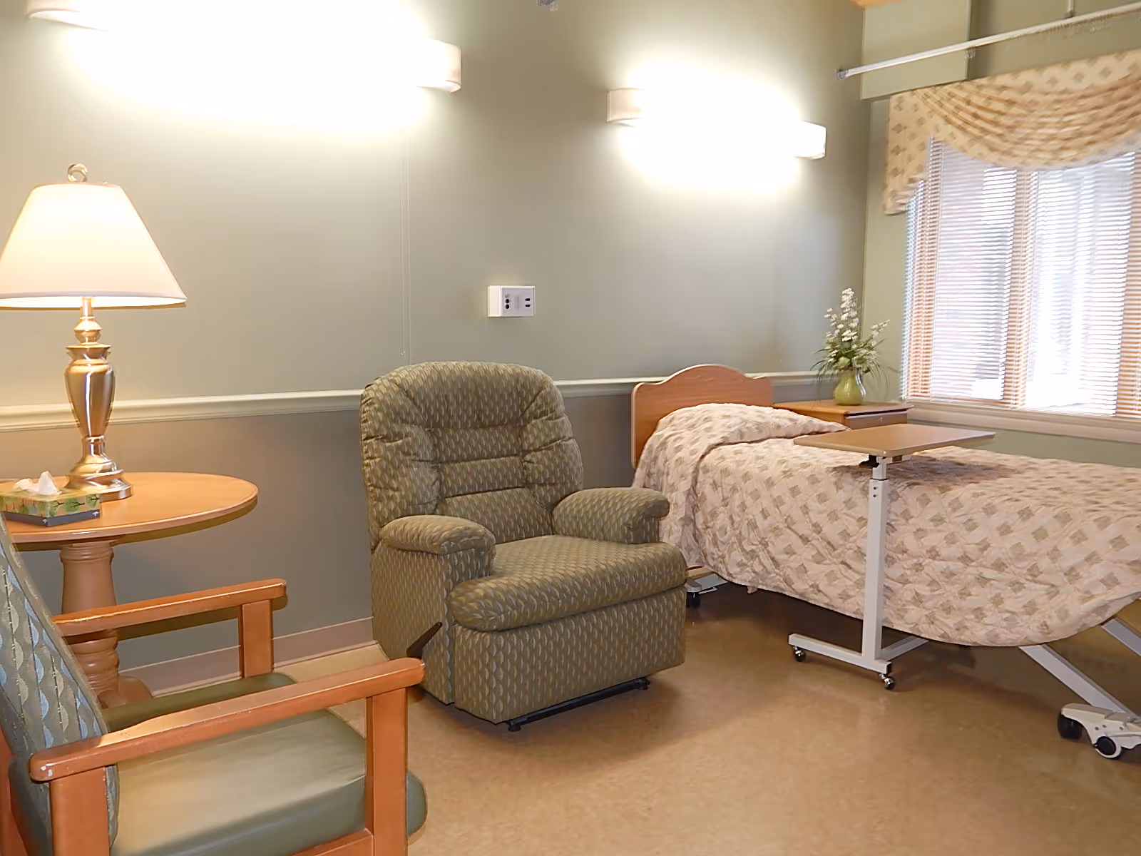 A cozy senior facility bedroom with a hospital-style bed, recliner, side table with lamp, and a window with blinds.