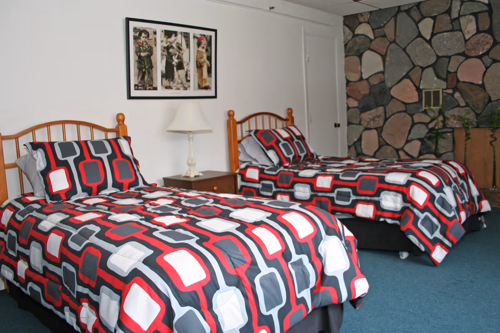 A bedroom with two twin beds featuring matching bedding with a geometric pattern in red, white, gray, and black. Between the beds is a wooden nightstand with a white lamp. The wall behind the beds is white with a framed picture of children, and one wall is made of large, irregularly shaped stones. The floor is carpeted in blue.