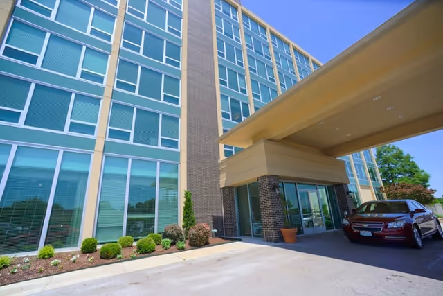 Exterior view of a multi-story building with large windows and a covered entrance. There are small bushes and plants along the building, and a red car is parked under the covered area. The sky is clear and blue.