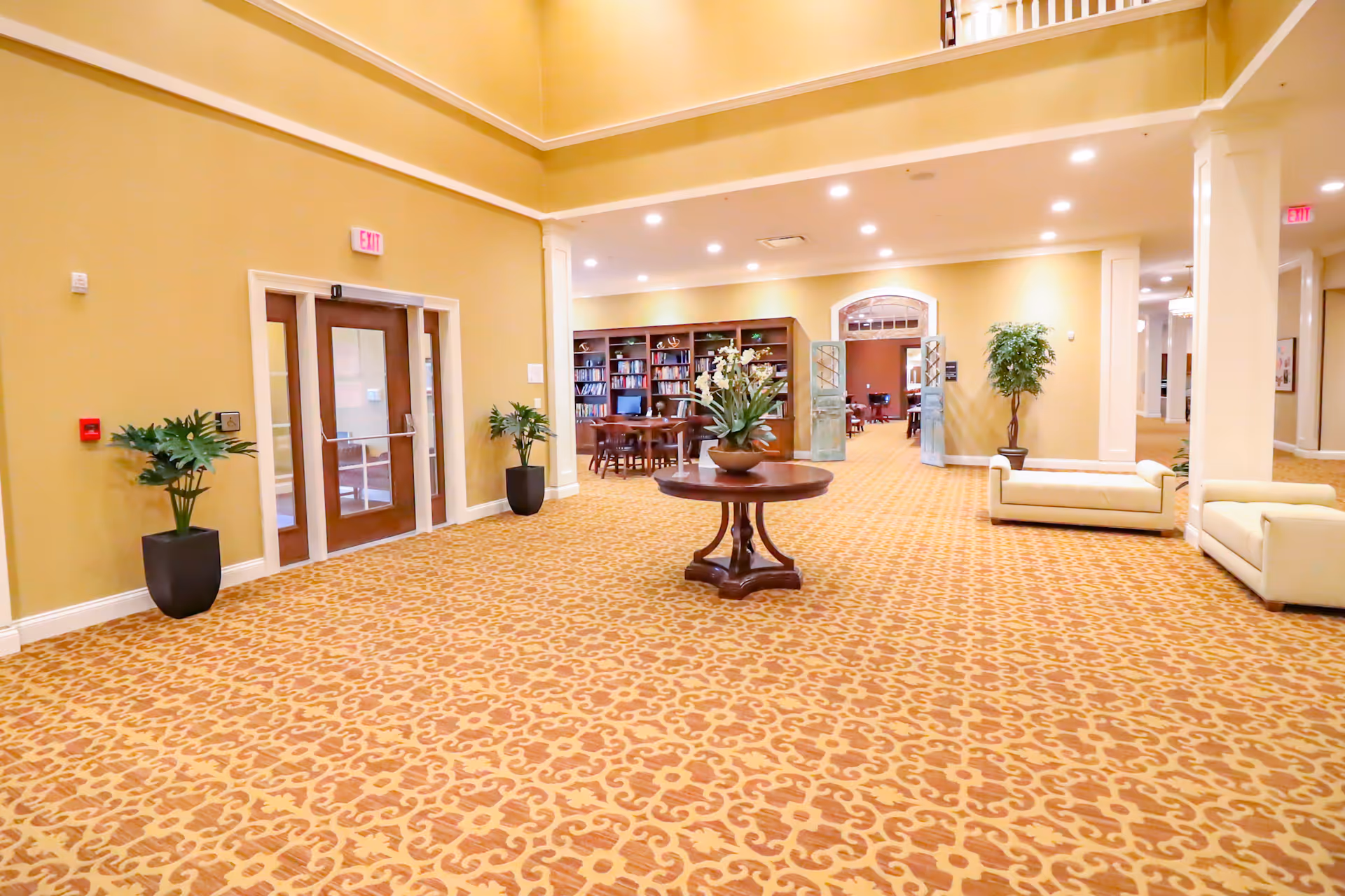 Spacious lobby/common area with patterned carpet, a central round table with flowers, seating alcoves, and a doorway leading to a library.