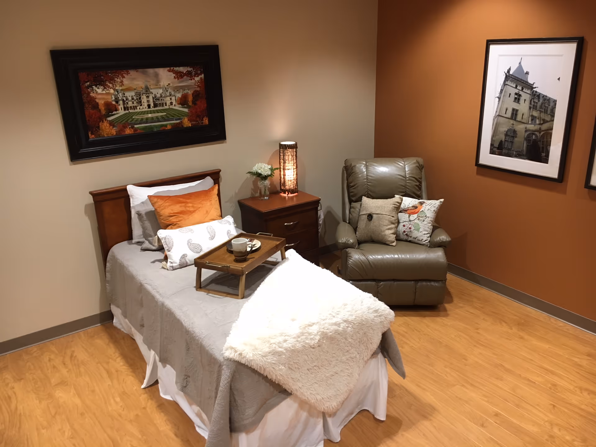 A cozy bedroom with a single bed covered in gray bedding and multiple pillows, including an orange one. A wooden tray with a cup and plate is placed on the bed. Next to the bed is a wooden nightstand with a lamp and a small vase of flowers. A comfortable gray recliner chair with decorative pillows is positioned in the corner. The walls are painted in beige and burnt orange, adorned with framed pictures of buildings. The floor is light wood.