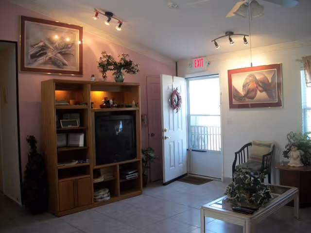 A cozy reception room with an entertainment center and TV, seating and coffee table, wall art, plants, and an open door beneath an EXIT sign.
