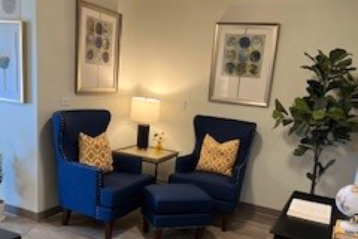 Two blue armchairs with a matching ottoman and a side table with a lamp sit against a wall with framed art and a potted plant.