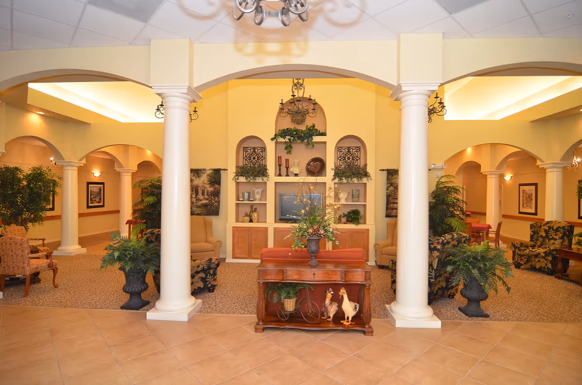 A spacious and warmly lit living room area in a senior living facility with cream-colored walls and large white columns. The room features comfortable armchairs and sofas arranged around a central wooden table with decorative plants and figurines. Built-in shelves with decorative items and a TV are set into the far wall, flanked by framed artwork and additional plants. The ceiling has recessed lighting and chandeliers, creating a cozy and inviting atmosphere.