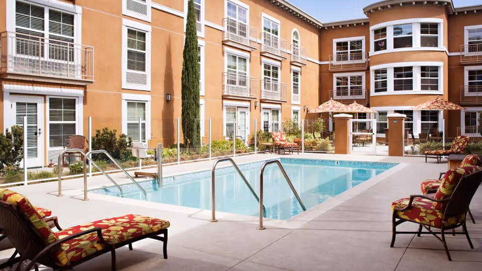 Outdoor swimming pool area at Belmont Village Senior Living Cardiff by the Sea with lounge chairs featuring red and yellow floral cushions, umbrellas, and a multi-story building with balconies and large windows surrounding the pool.
