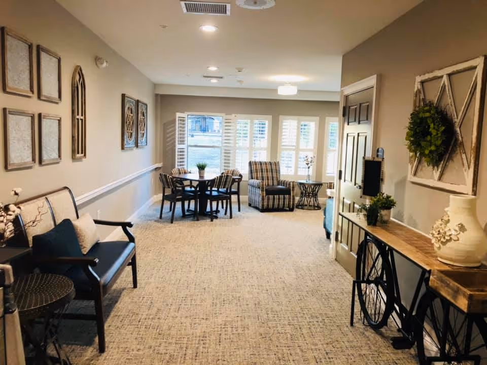 A cozy interior common area with beige walls and carpeted floor. The room features a bench with cushions on the left, a round table with four chairs near the windows, and a striped armchair with a small side table. Decorative wall art and plants adorn the space, creating a welcoming atmosphere.