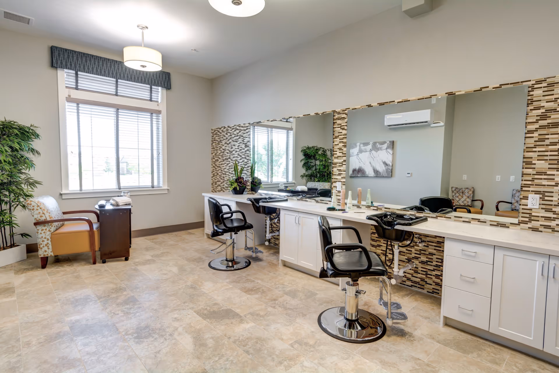 A bright and clean salon area with two black salon chairs in front of a long counter with large mirrors and hair care products. There is a patterned tile backsplash behind the counter. To the left, there is a window with blinds, a potted plant, and a cushioned armchair next to a small wooden side table. The floor is tiled and the walls are painted light gray.