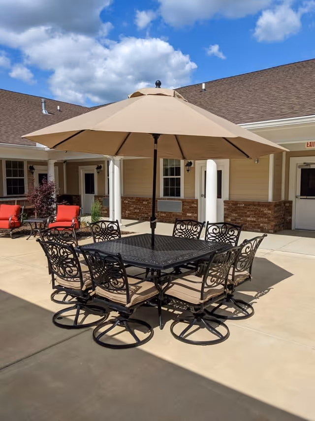 Outdoor patio area with a square black metal table surrounded by eight matching chairs with beige cushions. A large beige umbrella is centered over the table. In the background, there are red cushioned chairs, beige siding with brick accents, white columns, and a partly cloudy blue sky.
