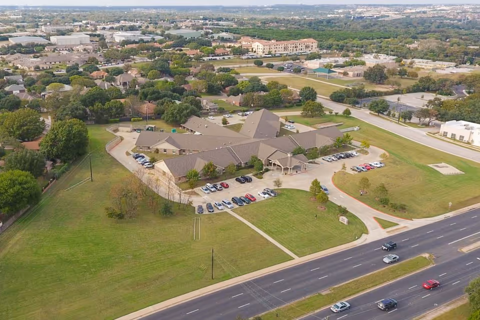 Aerial view of a single-story senior care facility with parking lots, green lawns, and surrounding neighborhood beside a multi-lane road.
