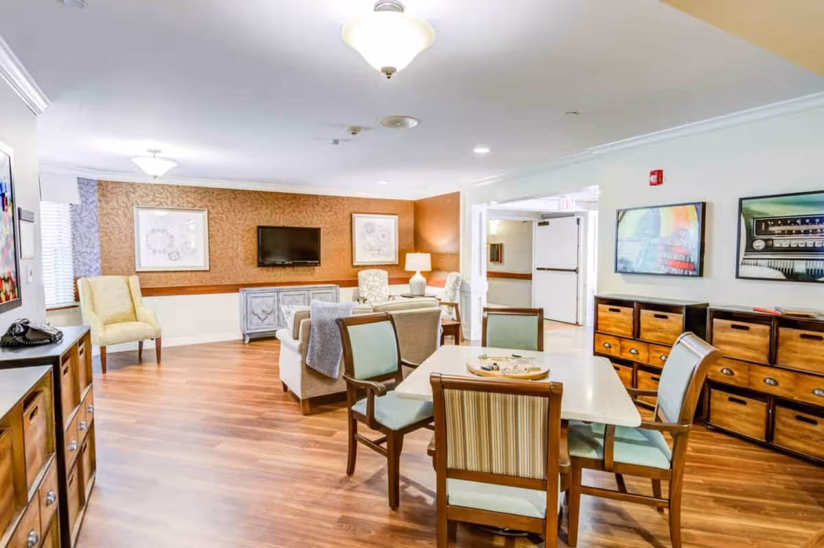 A bright and spacious common area in a senior living facility featuring a wooden floor, a table with four chairs, a seating area with sofas and armchairs, a wall-mounted TV, and decorative artwork on the walls.