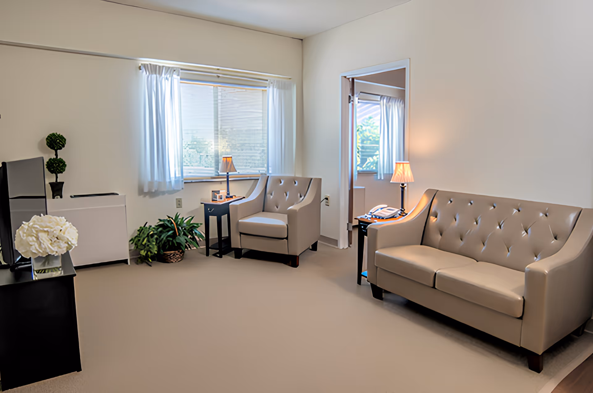 A bright and clean living room with beige leather sofa and armchair, two small black side tables with lamps, a window with white curtains, a potted plant on the floor, and a black TV stand with a white flower arrangement.
