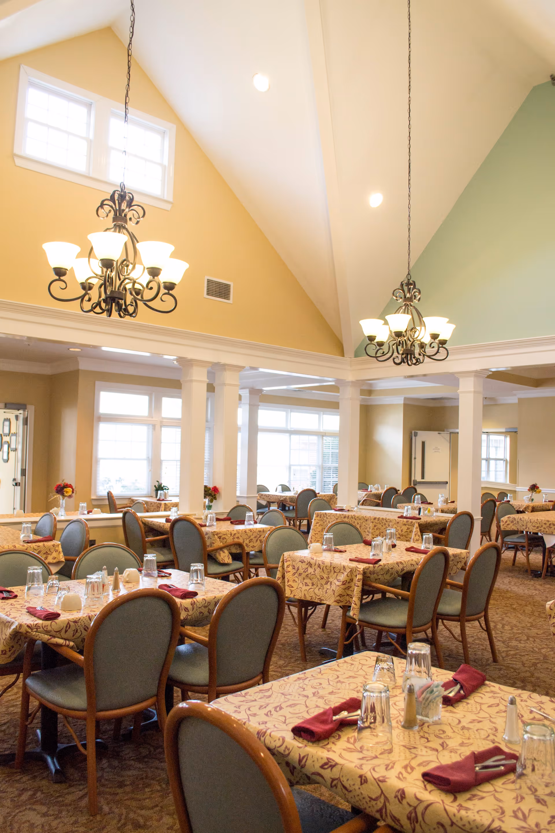 A bright and spacious dining room with multiple tables covered in patterned tablecloths, each set with glasses, napkins, and condiments. The room features high vaulted ceilings with two ornate chandeliers, large windows allowing natural light, and columns separating different sections of the room.