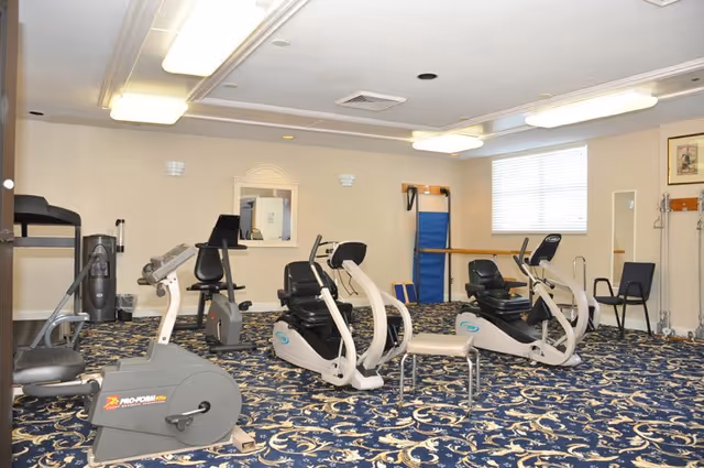 A fitness room with exercise equipment including stationary bikes and recumbent bikes on a patterned carpet. The room has beige walls, a window with blinds, fluorescent ceiling lights, a mirror, and some exercise mats and chairs along the walls.