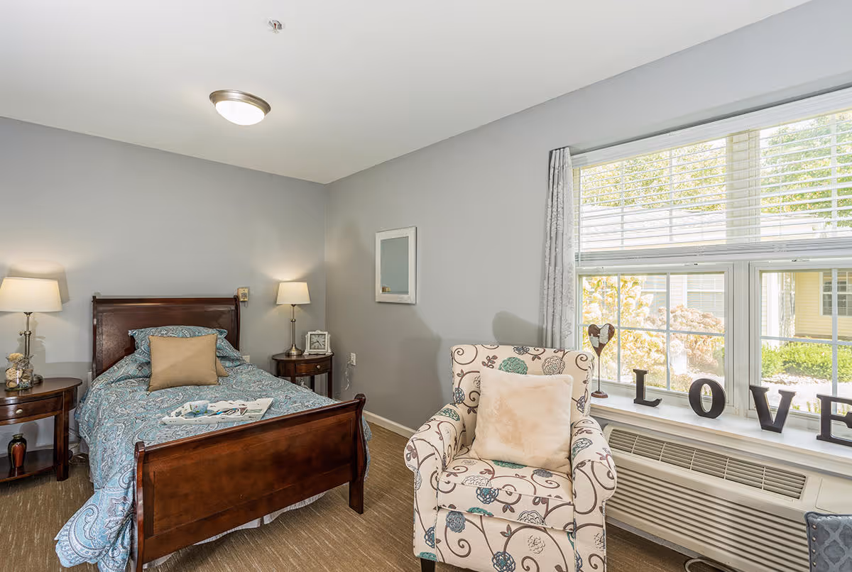 Furnished bedroom with a wooden bed, patterned armchair by a large window and decorative 'LOVE' letters on the sill.