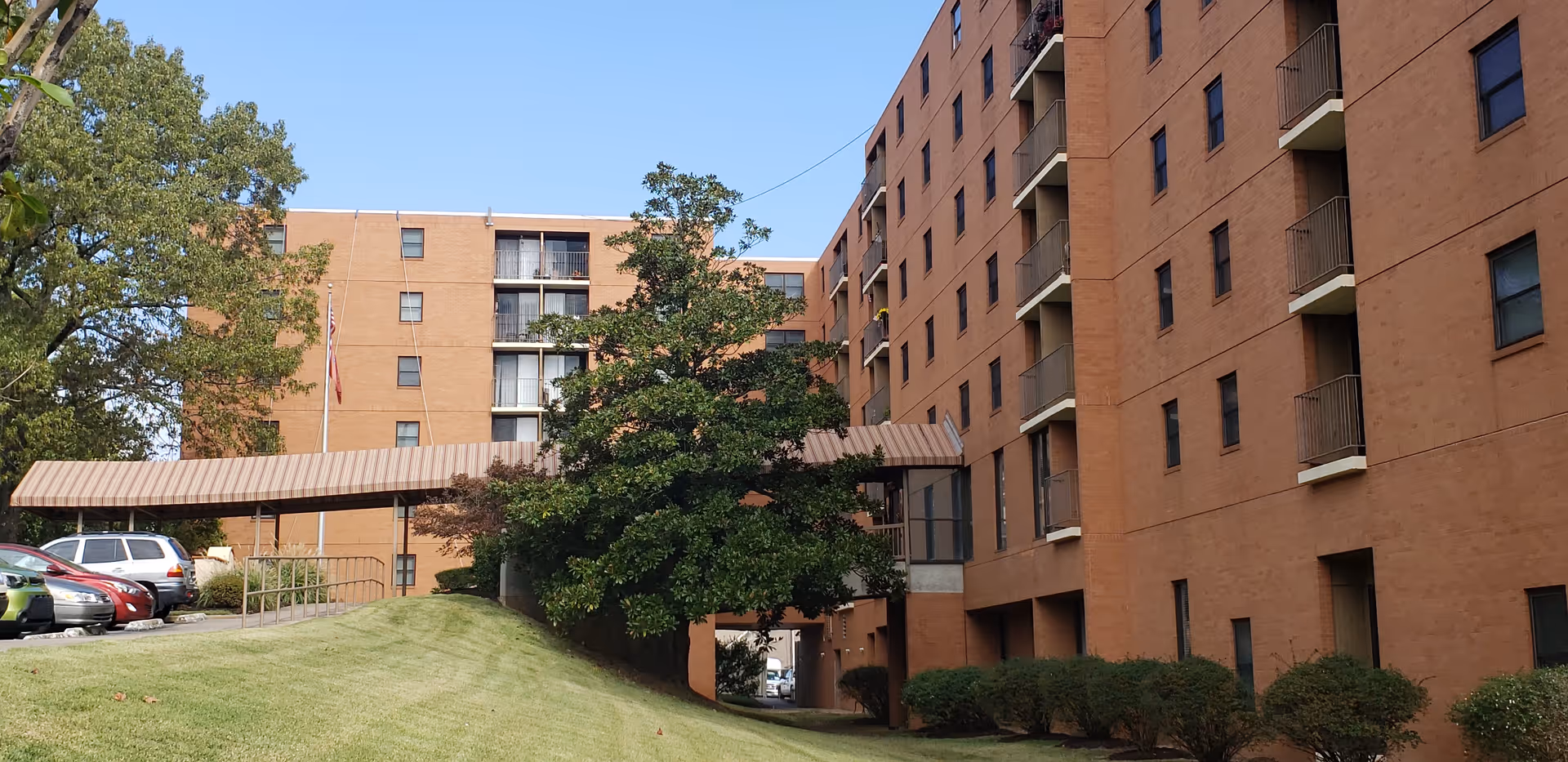 Exterior view of a multi-story brick building with balconies and windows, surrounded by green grass, trees, and parked cars under a clear blue sky.