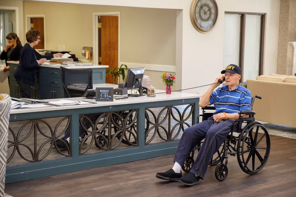 Elderly man in a wheelchair speaking on a telephone near a reception desk while staff work behind the counter in a senior living lobby.