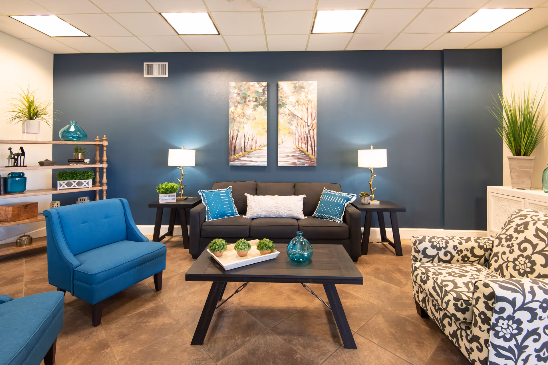 A cozy living room with a dark gray sofa adorned with blue and white pillows, two black side tables with lamps, a wooden coffee table with decorative plants and a blue glass vase, a blue armchair, a patterned armchair, and a wooden shelf with various decorative items against a blue accent wall with two nature-themed paintings.