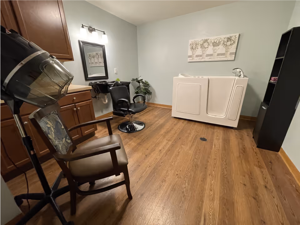 A room with wooden flooring featuring a hair dryer chair and a salon chair in front of a mirror and sink. There is a white walk-in bathtub against the far wall, a black shelving unit, and a decorative floral wall art above the bathtub.