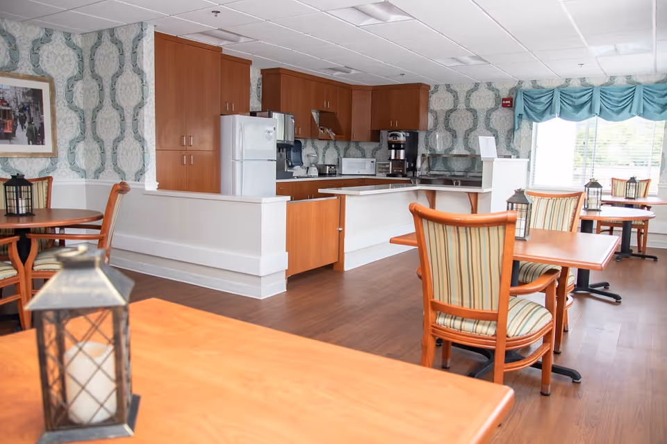 A bright dining area with wooden tables and striped cushioned chairs. The room features a kitchen area with wooden cabinets, a white refrigerator, microwave, coffee maker, and other appliances. The walls have patterned wallpaper, and there are large windows with blue valances letting in natural light. Lantern-style candle holders are placed on the tables.