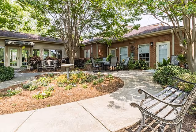 Outdoor courtyard area with paved walkways, garden beds with flowers and shrubs, several trees providing shade, metal benches, and a table with chairs. The courtyard is surrounded by a single-story brick building with windows and doors.