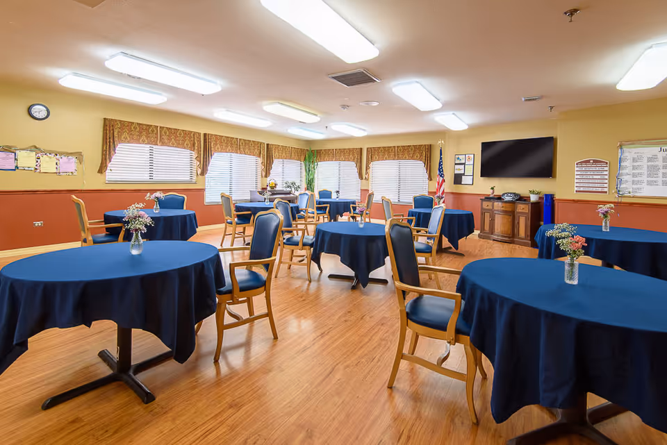 A bright dining room with multiple round tables covered in dark blue tablecloths, each adorned with a small vase of flowers. Wooden chairs with blue cushions surround the tables. The room has large windows with blinds and patterned valances, wood flooring, and fluorescent ceiling lights. An American flag stands in the corner near a wall-mounted TV and a wooden cabinet.