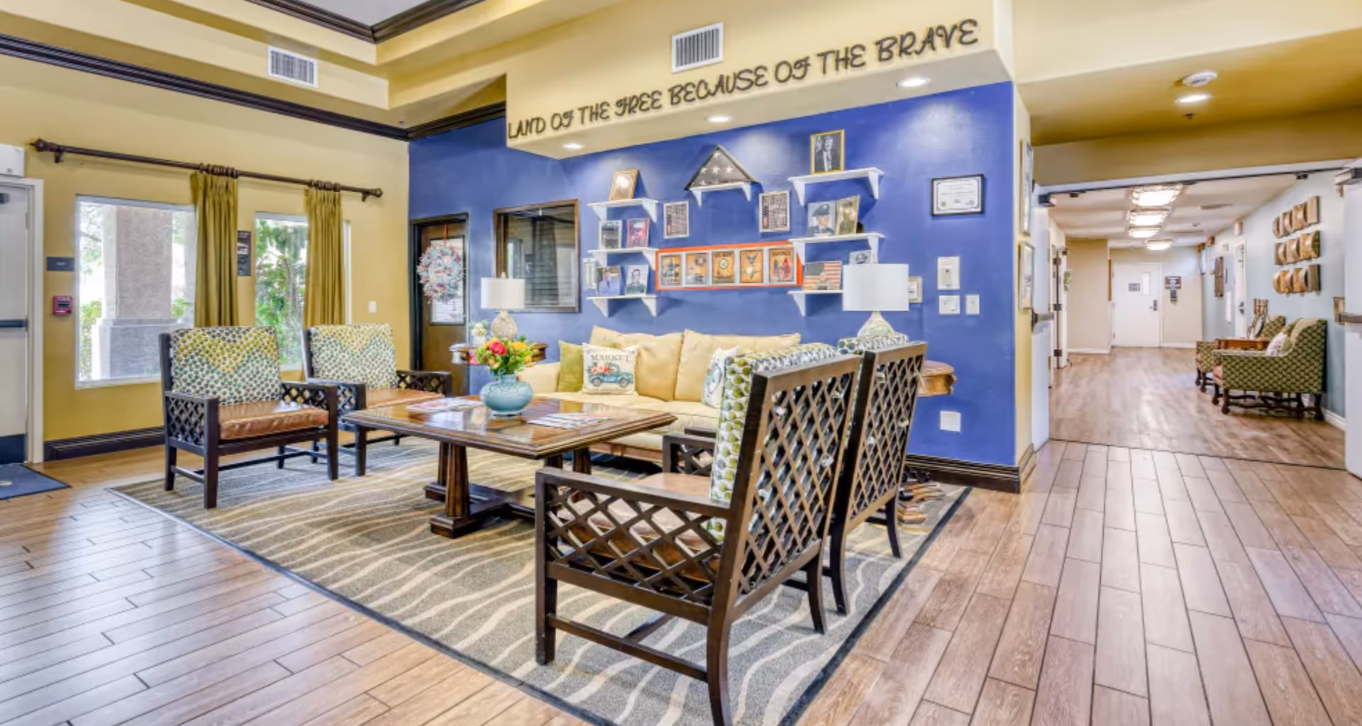A cozy seating area in a senior living facility with a beige couch, four wooden chairs with patterned cushions, and a wooden coffee table on a patterned rug. The wall behind the couch is painted blue and decorated with shelves holding framed photos, a folded American flag, and various memorabilia. Above the blue wall, the phrase 'LAND OF THE FREE BECAUSE OF THE BRAVE' is displayed. The room has wood-look flooring, yellow walls, and large windows with green curtains. A hallway with additional seating is visible to the right.