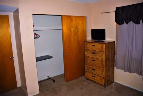 Small bedroom with dresser, TV, open closet, ironing board, and curtained window.
