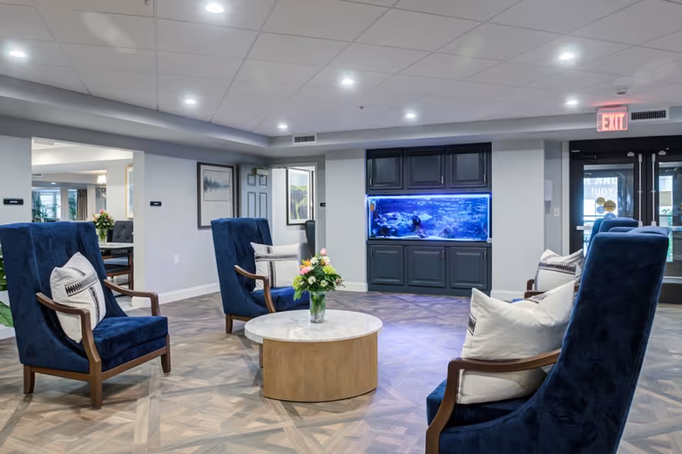 A bright seating area with blue upholstered armchairs arranged around a round coffee table and a built-in aquarium in the background.